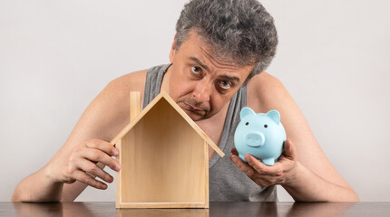 A middle-aged man in a sleeveless T-shirt with a piggy bank in his hands and a symbolic wooden house