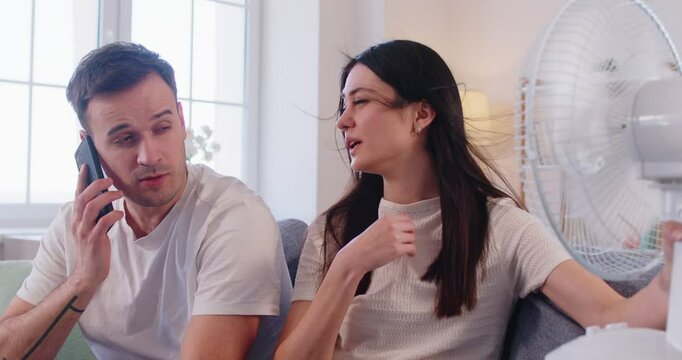 Family couple faces discomfort and trouble from hot summer weather at home living room, using fan for cool air in a challenging setting with focus on managing heat and relief every day.