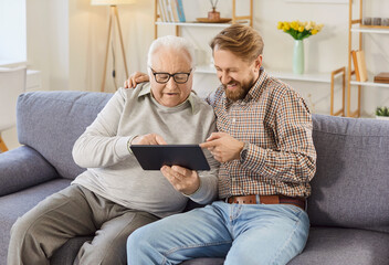 Portrait of elderly senior man sitting on sofa in living room at home with his adult son and using tablet together,surfing social media, e-banking, e-commerce or having online shopping on application