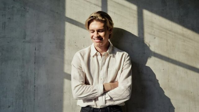 Happy young man smiling and leaning on a textured wall, wearing an unbuttoned long sleeve shirt with arms crossed in bright sunlight