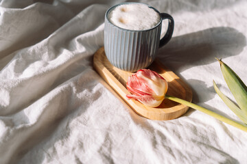 Coffee with foam on wooden stand next to tulip on white bed linen.
