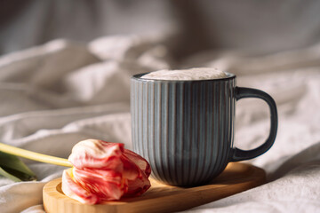 Coffee with foam on wooden stand next to tulip on white bed linen.