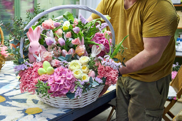 Close-up of a male florist in a yellow T-shirt holding a large Easter basket with pink flowers and...