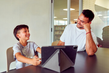 Happy teacher assisting young student with tablet and stylus in modern library setting, fostering...