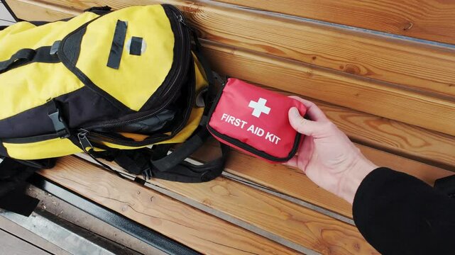 A person retrieves a stocked first aid kit from their yellow backpack while seated on a wooden bench, emphasizing the importance of preparedness for emergencies during outdoor activities
