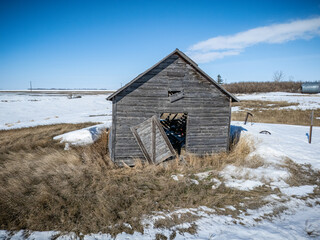 Small, old, wooden building with a broken door sits in a snowy field