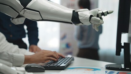 A robotic arm assists a human worker at a keyboard in a modern office, showcasing innovative technology and collaboration in a digital workspace environment.Noogenesis