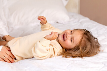 Hands of mom playfully tickles her happy laughing baby girl while she lying on white bed in a...