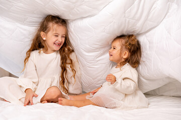 Children sisters in bed under the blanket having fun, smiling. Happy little girl and her newborn baby sister hiding under the white blanket. Childhood concept. Copy space. High quality photo
