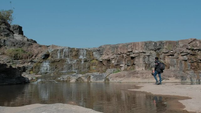 South Asian model posing near a cliffside waterfall in a rocky landscape surrounded by nature
