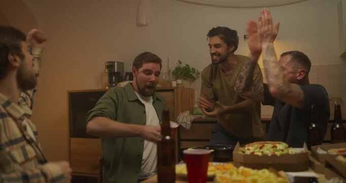 A brunette guy finishes the whole glass to the bottom and his friends support him and applaud during a cheerful bachelor party at home in the evening in the kitchen