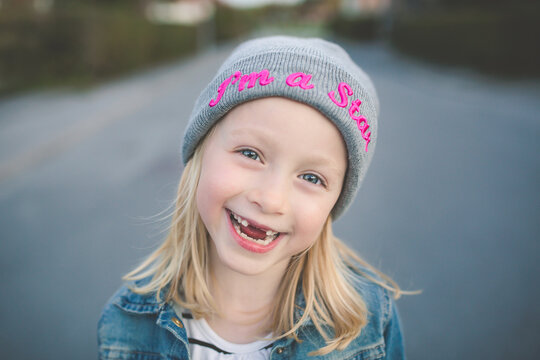 Close-up portrait of a Smiling  girl with her front teeth missing standing in the street wearing a beanie with the words I'm a star