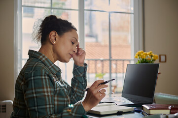 Focused Woman Working Remotely at Home With a Laptop and Notebook