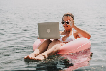 A woman is floating in a pink inflatable raft with a laptop in her hand. She is smiling and she is...