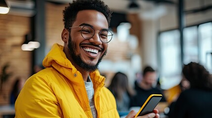 Obraz premium A smiling man with a beard stands in a modern office environment, wearing a tan jacket over a white shirt, holding a smartphone in his hands.