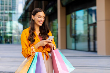 Message about discount. Cheerful young woman with colored shopping bags typing in smartphone...