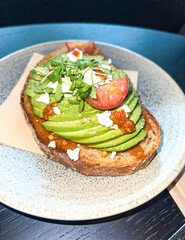 Avocado toast bread, fruit slices garnished with tomatoes and feta cheese, microgreens, in ceramic plate on round black table in restaurant, high angle view, vertical shot, copy space