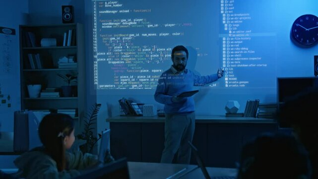 Full shot of mature male coding teacher explaining new computer algorithm and pointing at variables for schoolkids with laptops during lesson, in dark classroom and blue projector light