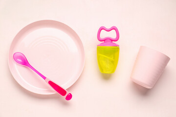 Plate with spoon, cup and nibbler for baby on pink background
