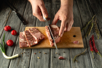 Skillful hands are slicing cooked meat on a wooden board, surrounded by fresh garlic, herbs, and vegetables, showcasing culinary preparation in a rustic kitchen