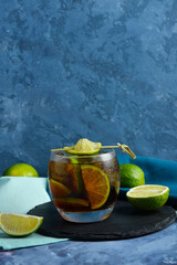 Board with glass of cold Cuba Libre cocktail and limes on blue background