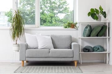 Stylish grey sofa, shelving unit with pillows and houseplants near window in living room