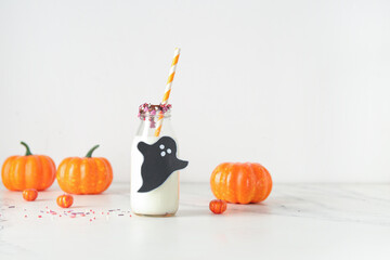 Bottle of milk, ghost made of paper, sprinkles and pumpkins for Halloween on light background