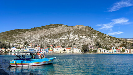 Fototapeta premium Colorful Waterfront Houses And Blue Fishing Boat In A Calm Bay, Peaceful Coastal Town At The Foot Of A Rocky Hill, Bright Sunny Day, Seaside Tranquility, Mediterranean Island Life