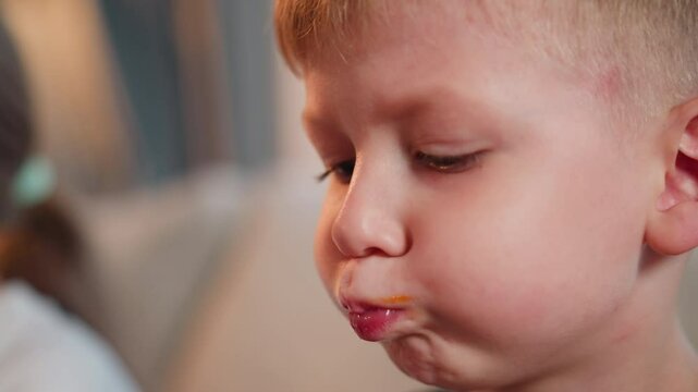 close up of young boy with cheeks puffed out savoring toasted bread with sauce stain on lips as he licks mouth beside blurred figure of girl with ponytail seated near him