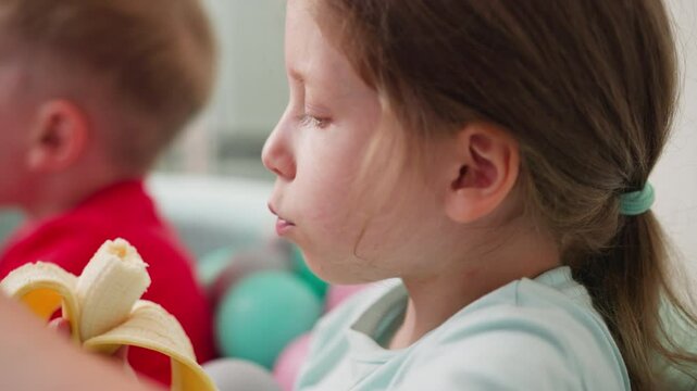 Close up side view of little girl peeling and eating nearly finished banana while sitting in pastel ball pit, brother nearby slightly blurred in background