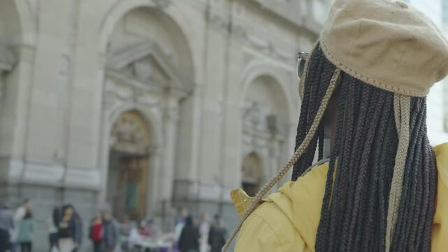 Black Latina woman with braids, yellow jacket, beret, and sunglasses, standing amazed as the camera circles 360&deg; around her, sightseeing in a big city with a mix of modern and classical architecture