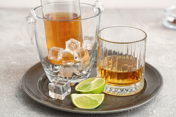 Bottle and glass of cold rum with ice cubes and lime on plate on table against grey background. Closeup