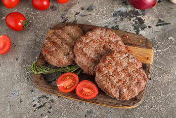 Wooden board with tasty meat cutlets, tomatoes and rosemary on grey grunge background