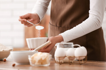 Woman adding sugar into bowl for preparing chocolate brownie at table in kitchen. Closeup