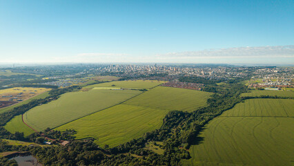 Aerial view of Cascavel, Paraná, Brazil, with large agricultural fields and forested areas in the foreground and the city skyline in the distance.