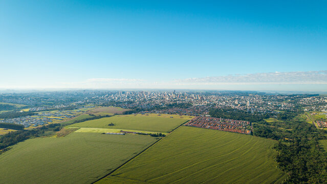Wide aerial view of Cascavel, Paran&aacute;, Brazil, with agricultural fields and forest in the foreground and a dense urban skyline in the background.