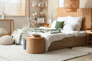 Interior of bedroom with big bed, shelving unit, wicker table and blooming branches in vases