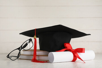 Mortar board with books, diploma and eyeglasses on white wooden background