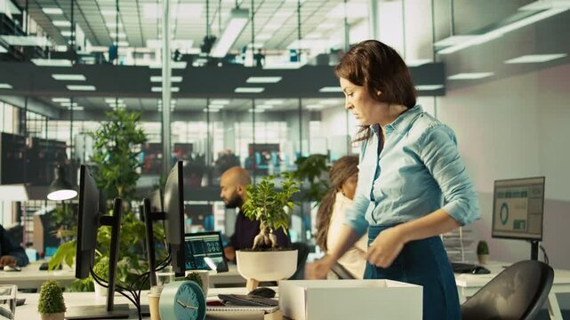 Upset worker in office packing desk plants on last day at work after quitting, saying goodbye to colleague. Employee getting fired from job during layoffs, saddening coworkers, camera B