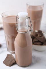 Tasty chocolate milk in bottle, glasses and pieces of chocolate on white marble table, closeup