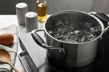 Cooking pot with boiling water and stove on light table against grey background, closeup