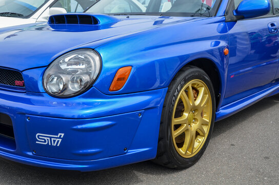 Close-up shot of the front of a blue Subaru Impreza WRX STI. The car features a distinctive hood scoop, gold-colored wheels, and the "STI" logo on the front bumper 