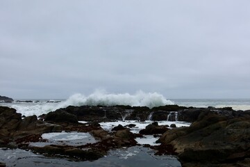 Ocean waves crash against rugged rocks, sending white spray into the air along a dramatic coastal shore.