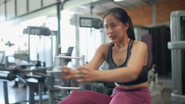 Dedicated asian woman performing seated cable rows with intense focus, targeting back muscles and demonstrating strength training commitment in contemporary fitness facility