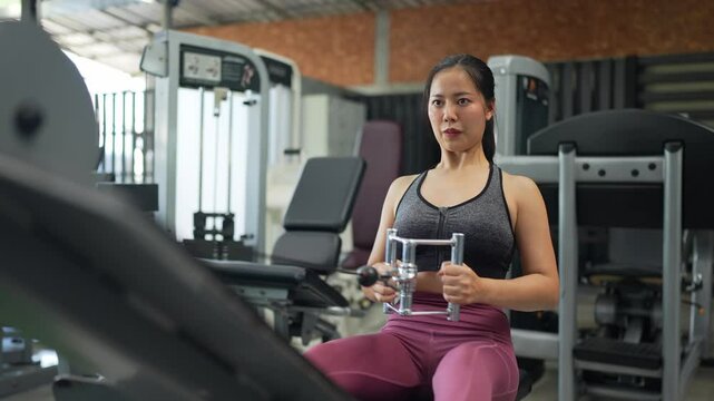 Focused asian woman in activewear strengthens back muscles with seated cable rows, demonstrating proper form and controlled movements in a modern fitness center