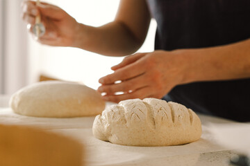 Woman hands skillfully score the surface of a round dough loaf, preparing sourdough bread for baking. The bright kitchen setting provides a warm atmosphere and fresh ingredients.
