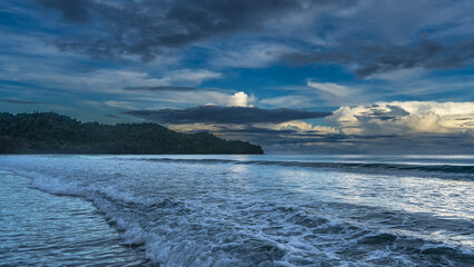 Morning by the ocean. The waves roll towards the shore, foam, and spread out. The clouds in the blue sky are highlighted in gold. A hill in the distance. Malaysia. Borneo. Kota Kinabalu.