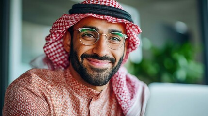 Smiling arab man wearing traditional keffiyeh headdress looks at the camera with round glasses. The man has a groomed beard.