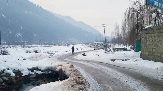 Man walking along a snowy road with the majestic Himalaya mountains in the distance and frozen lidder river on the right showing the beautiful sights of Pahalgam