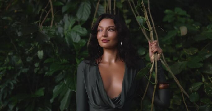 Sexy and confident Caucasian woman in a deep-cut green bodysuit stands surrounded by tropical jungle vines. This high-quality conceptual footage is ideal for beauty, fashion, or promo use.